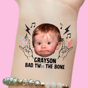 May include: A temporary tattoo featuring a baby's face with the name "Grayson" and the phrase "Bad to the Bone." The design includes rock and roll hand gestures, musical notes, and a skull and crossbones.