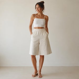 Könnte beinhalten: Ein weißes bauchfreies Top mit dünnen Trägern kombiniert mit knielangen, weit geschnittenen Shorts in Off-White mit elastischem Bund und Seitentaschen. Das Outfit wird durch schlichte, helle Zehentrenner-Sandalen abgerundet.