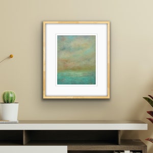Puede incluir: Impresi&oacute;n art&iacute;stica enmarcada de un paisaje abstracto. La obra presenta una escena serena con una zona de agua turquesa y verde azulado, un horizonte naranja y marr&oacute;n apagado y un cielo nublado en tonos azules y amarillos. La impresi&oacute;n est&aacute; enmarcada en oro con un passe-partout blanco.