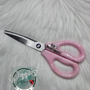 May include: A pair of scissors with silver metal blades and pink glitter handles. The blades are marked with the text CHNZAO and the handles feature the black text Mrs. Rodriguez. The scissors have a central screw and two finger loops.