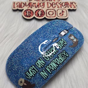 May include: A blue glitter computer mouse featuring a graphic of two brown otters and the text "JUST AN OTTER DAY IN PARADISE" in black lettering. Above the mouse is a wooden sign with the text "DAZZLING LADYBUG DESIGNS" and social media icons.