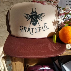 May include: A beige and maroon trucker hat with a black bee graphic and the word "Grateful" printed on the front. The hat has a mesh back and a curved brim. A sprig of white flowers and an orange are in the background.