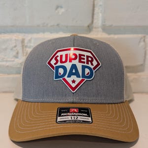 May include: A gray and tan trucker hat with the words "SUPER DAD" in a red, white, and blue shield design. The hat has a curved brim and a mesh back. The Richardson brand label is visible.