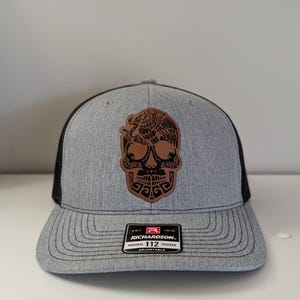 May include: A gray and black trucker hat with a brown leather patch featuring a black skull with an eagle design. The patch has the text "EST. 1970 RICHARDSON. ORIGINAL 112 TRUCKER ADJUSTABLE".