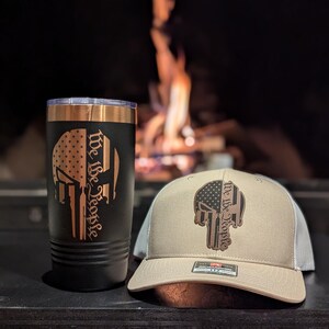 May include: A black and copper travel mug with a skull graphic and the text "We the People" on the side. A tan baseball cap with a similar skull graphic and text is also shown.