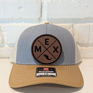 May include: A trucker hat with a light blue crown, tan brim, and white mesh back. A circular brown patch features the letters "M", "E", and "X" with a fish graphic. The hat includes a "RICHARDSON" tag.