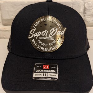 May include: Black trucker hat with a gold metal patch that says "I can do all things Super Dad Philippians 4:13 Through Christ who strengthens me". The hat has a black mesh back and a Richardson 112 label.