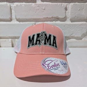 May include: Pink and white baseball cap with a black and white "MAMA" patch with a lightning bolt and leopard print. The cap has a white mesh back and a black "Evolve Eludes" tag with the text "Concealed Ponytail Opening".