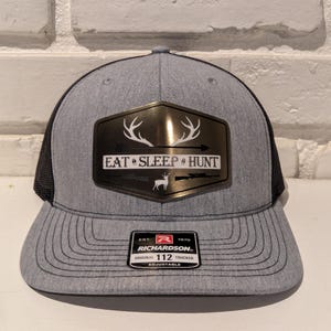 May include: A gray and black trucker hat with a gold metal patch that says "Eat Sleep Hunt". The patch features a deer head with antlers and an arrow. The hat is adjustable and has a Richardson 112 label.