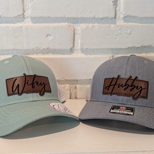 May include: Two trucker hats, one mint green and one gray, with white mesh backs. The mint green hat says "Wifey EST 2025" on a brown leather patch. The gray hat says "Hubby EST 2025" on a brown leather patch.