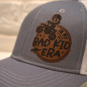 May include: Gray and white trucker hat with a brown leather patch that says "In My Bad Kid Era" with a cartoon of a person riding a dirt bike.