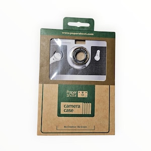 May include: A camera case in a cardboard box with a clear window. The camera is silver and black with a lens and a dial. The box has the text "camera case" and "Be Creative - Be Green". The website address www.papershoot.com is printed on the top.