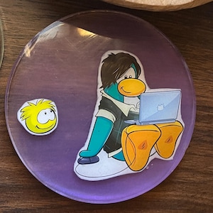 May include: A round, purple coaster featuring a cartoon penguin with a laptop and a yellow character. The penguin is wearing a black shirt and orange flippers. A small yellow character is to the left of the penguin.