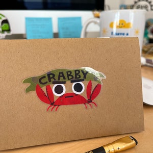 May include: A brown card with a crab sticker that says "CRABBY". The crab is red with white eyes and green seaweed on its head. A black Palomino Blackwing pencil with gold accents is in the foreground.