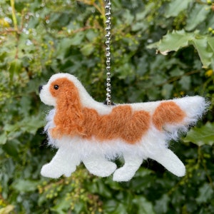 May include: A small, felt Cavalier King Charles Spaniel dog keychain. The dog is brown and white with a long, flowing tail. The keychain is attached to a silver chain.