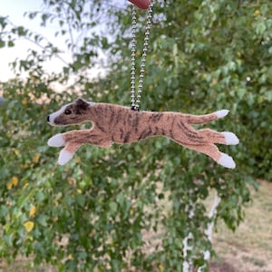 May include: A felt ornament of a running dog, suspended by a silver chain. The dog has a brown and tan brindle pattern with white accents on its paws and snout. The background is blurred green foliage.