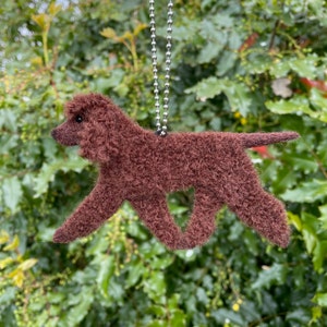 May include: A brown felt dog ornament hanging from a silver chain. The dog is in a running pose with its tail curled up.