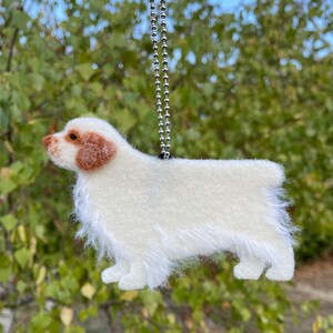 May include: A white felt ornament of a Clumber Spaniel dog hanging from a silver chain. The dog has brown ears and a fluffy white tail.