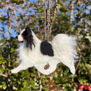 May include: A handmade, white and black Papillon dog ornament. The dog has a black spot on its back and black and brown markings on its head. It is suspended by a silver chain, perfect for hanging as a decoration.
