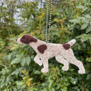 May include: A brown and white felt dog ornament hanging from a silver chain. The dog is in a running pose with its tongue out. The ornament is detailed with brown spots and a brown nose.