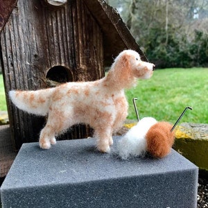 May include: A needle felted dog sculpture in a light brown and white color. The dog is standing on a gray foam block with a ball of white and brown wool yarn next to it. The background is a wooden birdhouse and green grass.