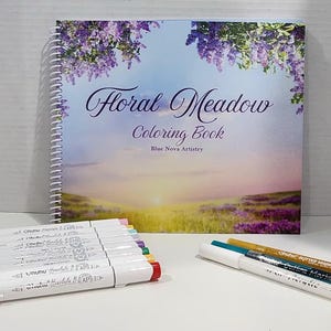 May include: A spiral-bound "Floral Meadow Coloring Book" with a landscape cover featuring purple flowers and a sunset. Several white markers with colorful tips and two glitter markers are in front of the book.