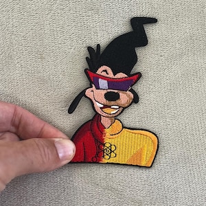 May include: Embroidered patch featuring a cartoon character with black hair, a black hat, and red-purple sunglasses. The character wears a red and yellow jacket with a floral design. The patch is held by a hand.