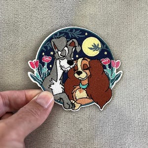 Op de afbeelding: Geborduurde patch met twee honden, een grijze en een bruine, in een romantische omhelzing. De patch heeft een donkerblauwe achtergrond met een volle maan en bloemaccenten. De patch wordt door een hand vastgehouden.