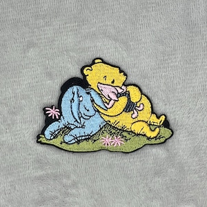 May include: Embroidered patch featuring Winnie the Pooh and Piglet hugging, with blue and pink accents. The patch has a black border and is set on a green grassy area with pink flowers. The characters are yellow and pink.