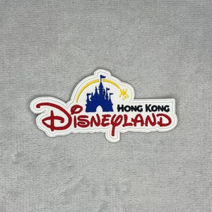 Può includere: Una toppa ricamata con il logo di Hong Kong Disneyland. Il design include la sagoma di un castello blu, una scia gialla ad arco con una stella, la scritta Disneyland in rosso e il testo Hong Kong in nero sulla destra.
