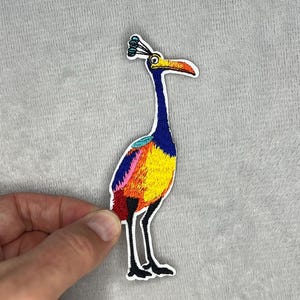 Puede incluir: Parche bordado con un pájaro colorido con cuello y pico largos. El pájaro tiene cuello azul, cuerpo amarillo y pecho rojo, naranja y rosa. El parche está delineado en blanco y tiene patas negras.