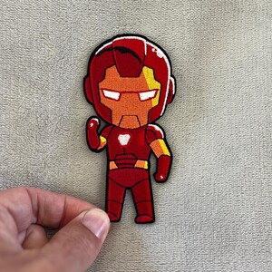 May include: Embroidered patch featuring a cartoon Iron Man character in a standing pose. The patch has a red, orange, yellow, and white color scheme with black outlining. The Iron Man patch is a superhero-themed item.