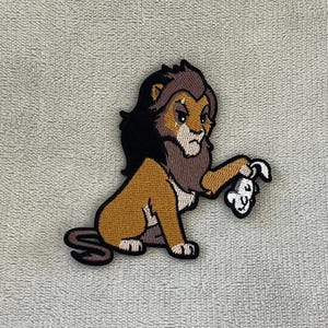 Peut inclure: Écusson brodé représentant un lion de dessin animé avec une crinière marron foncé et un corps beige. Le lion tient un os blanc dans sa patte. Le patch a des coutures noires et blanches.