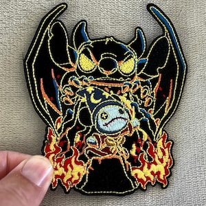May include: Embroidered patch featuring a stylized Stitch character with a devilish design. The patch is primarily black with yellow, orange, and blue accents, and a fiery border. The patch is held by a hand.