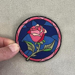 May include: Embroidered patch featuring a pink rose with green leaves on a stem, set against a stained-glass style blue background. The rose is framed by a pink border and a black outline. The patch is round.