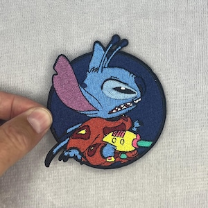 May include: A circular embroidered patch featuring a blue alien character with large ears and antennae. The character has a grumpy expression, wears a red garment, and holds a yellow and green ray gun against a dark blue circular background.