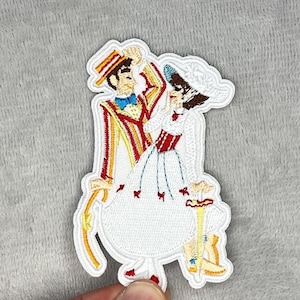 May include: Embroidered patch featuring a couple in vintage attire, likely from a musical. The man wears a striped suit with a bow tie and hat. The woman is in a white dress and hat. The patch has a white border.