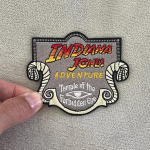 May include: Embroidered patch featuring the text "Indiana Jones! Adventure" in red and yellow, with "Temple of the Forbidden Eye" below. The patch has a gray background with a decorative border and two snake-like designs.