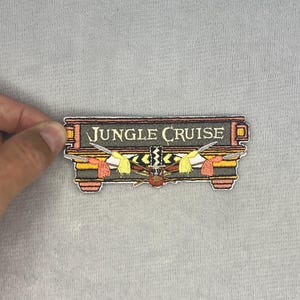 May include: A rectangular embroidered patch featuring the text JUNGLE CRUISE in white lettering on a dark grey background. The design includes a central tribal mask motif with yellow and orange accents, flanked by crossed spears and decorative elements.