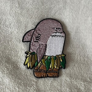 Puede incluir: Parche bordado para planchar de un tiburón de dibujos animados con un collar de flores y sandalias. El tiburón tiene una expresión gruñona y es de color gris, blanco y marrón.