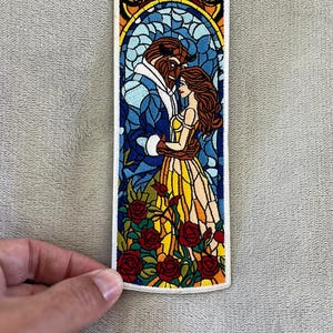 以下が含まれることがあります： 美女と野獣のステンドグラスデザインの刺繍パッチ。パッチは、バラに囲まれたキャラクターが抱き合っている様子を描いています。カラーパレットには、青、黄、赤、茶色が含まれています。