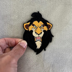 May include: Embroidered patch depicting a lion's head with a black mane, yellow and brown details, and a fierce expression. The patch is intricately designed and suitable for application on garments or accessories.
