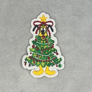 Puede incluir: Un parche bordado que muestra un personaje de perro de dibujos animados con pelaje amarillo y orejas negras, disfrazado de árbol de Navidad. El diseño incluye una estrella amarilla en la punta, un lazo rojo, ramas verdes y adornos rojos.