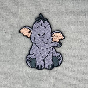 Puede incluir: Un parche bordado que muestra un elefante de dibujos animados con piel de color morado claro, orejas grandes de color melocotón y pies de color gris oscuro. El elefante está sentado con un pequeño mechón de pelo y un borde negro.