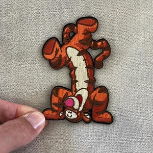 Könnte beinhalten: Ein gestickter Aufnäher mit Tigger aus Winnie Puuh. Der Aufnäher zeigt Tigger kopfüber mit Armen und Beinen in der Luft. Der Aufnäher ist orange mit schwarzen Streifen und einem weißen Bauch.