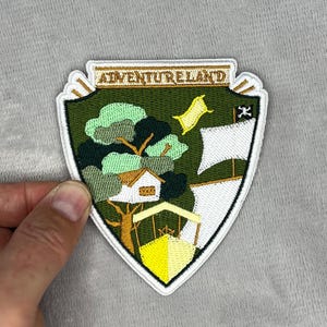 May include: Embroidered Adventureland patch with a shield design. The patch features a green background with a treehouse, a boat, and a yellow carpet. The text "ADVENTURELAND" is at the top.
