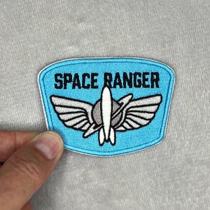 以下が含まれることがあります： 白い縁取りが施された水色の刺繍ワッペン。黒い文字で「SPACE RANGER」と書かれており、その下には白いロケットのグラフィックと、デザインの中央に配置された円形の惑星要素、そして様式化された翼が描かれています。