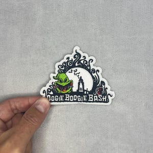 Può includere: Una toppa ricamata raffigurante un personaggio verde con un ampio sorriso, una luna piena bianca, la sagoma scura di una torre e motivi neri vorticosi. Il testo OOGIE BOOGIE BASH è ricamato in bianco nella parte inferiore su sfondo nero.