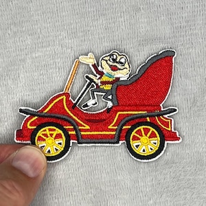 May include: Embroidered patch featuring a cartoon frog driving a red vintage car. The car has yellow wheels and gold accents. The frog is wearing glasses and a suit, waving from the driver's seat. The patch has a white border.