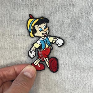 Peut inclure: Écusson brodé représentant Pinocchio du film Disney. Le patch montre Pinocchio dans sa tenue classique : un chapeau jaune avec une bande bleue, des gants blancs, une chemise blanche, un short rouge et des chaussures rouges. Le patch est sur un fond gris.